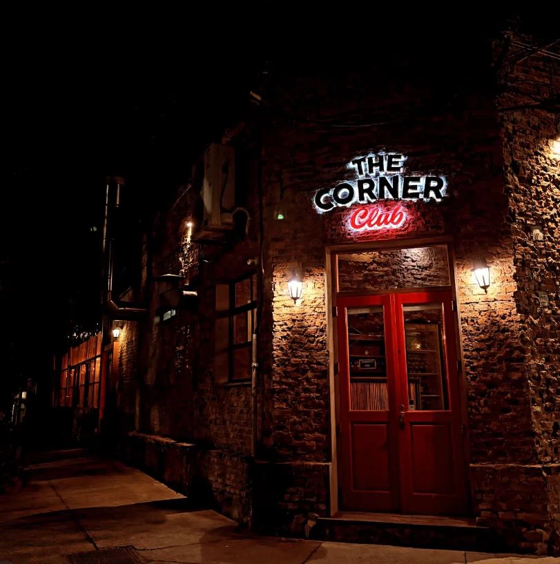 Frente de The Corner Club de noche: pared de ladrillo, cartel de neón y la puerta roja iluminada por dos faroles.