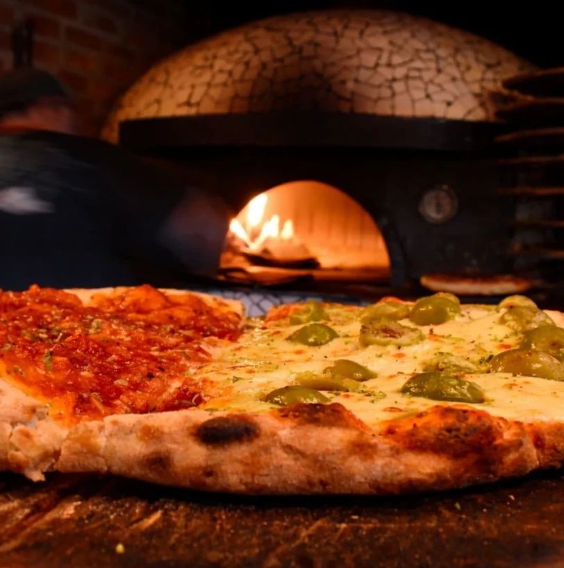 Pizza mitad muzzarella con aceitunas y mitad con salsa, frente al horno de leña encendido.